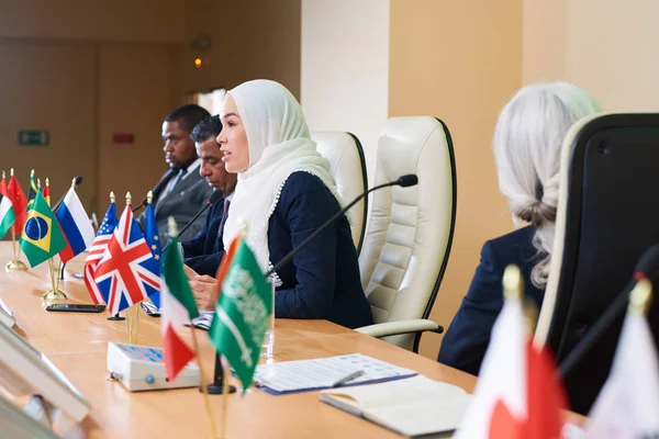 Confident young muslim female speaker talking in microphone at conference or political forum among foreign colleagues