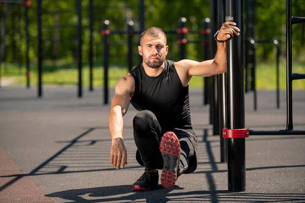 Young muscular athlete sitting on squats with left leg stretched forwards and holding by sports bar on outdoor sportsground