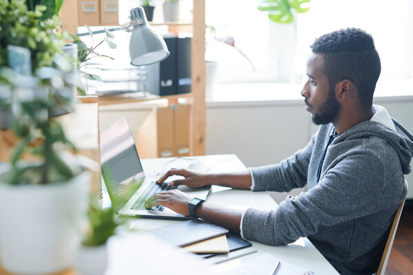 Young serious creative man in casualwear looking at laptop display while typing on keypad by his desk in office