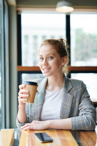 Portrait of positive beautiful redhead student girl in checkered jacket sitting at table and drinking coffee in fast food restaurant