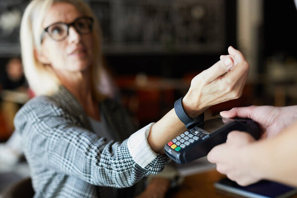 Modern blonde female keeping her wrist over electronic payment machine while paying for order through smartwatch in cafe