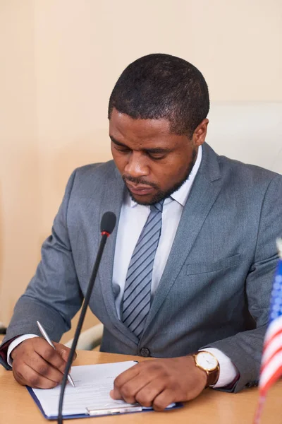 African-american businessman or delegate in suit sitting by table in front of microphone and looking through text of his report