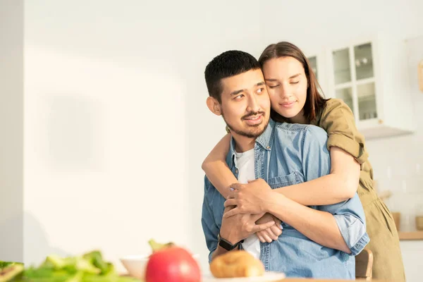 朝食に新鮮な野菜を入れた台所のテーブルのそばに立っている間 若い不愉美な女性が夫を抱きしめる — ストック写真