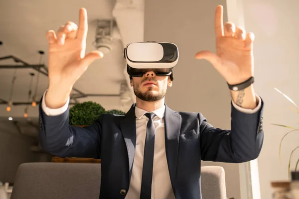Elegant businessman in formalwear and vr headset sitting in cafe while touching virtual display during presentation