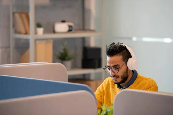 Serious thoughtful young Arabian office worker in eyeglasses sitting at table with partitions and listening to audio file in headphones while working in office