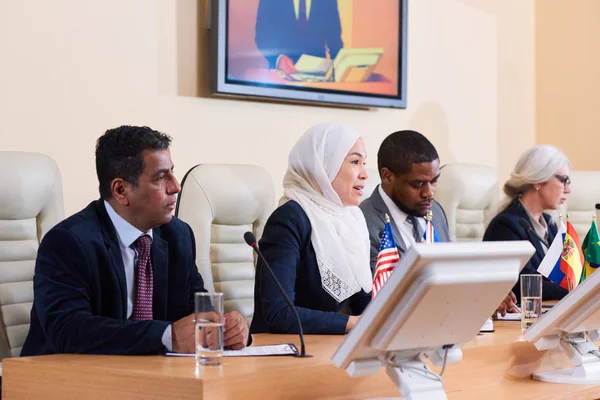 Young Muslim woman in white hijab making speech at business or political conference among foreign colleagues