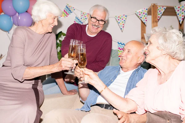 Cheerful friendly seniors clinking with flutes of champagne while having good time at home party