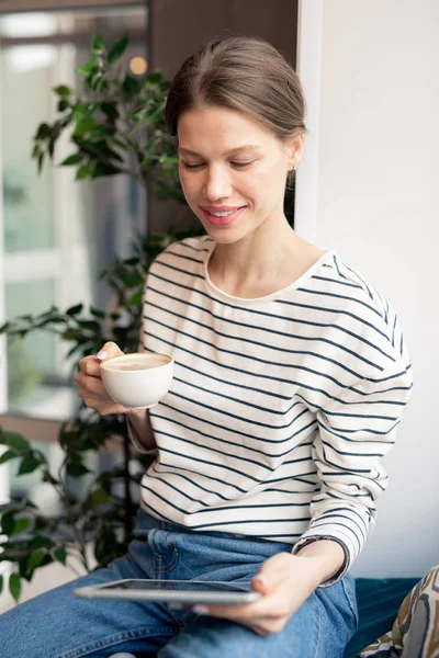 青いジーンズとストライプのプルオーバーでコーヒーを飲み カフェのタッチパッドでビデオを見ているかわいい若い笑顔の女性 — ストック写真