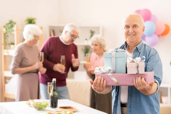 Senior smiling man with several packed gifts looking at you at home party with friends