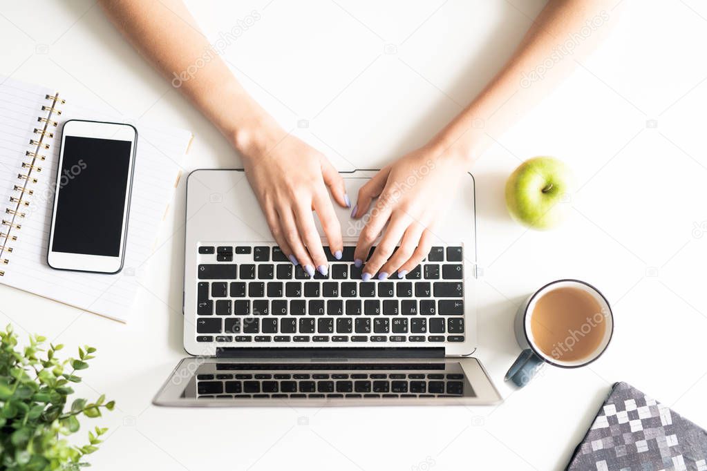 Overview of human hands on laptop keys surrounded by mug with coffee, green apple, smartphone and open notebook