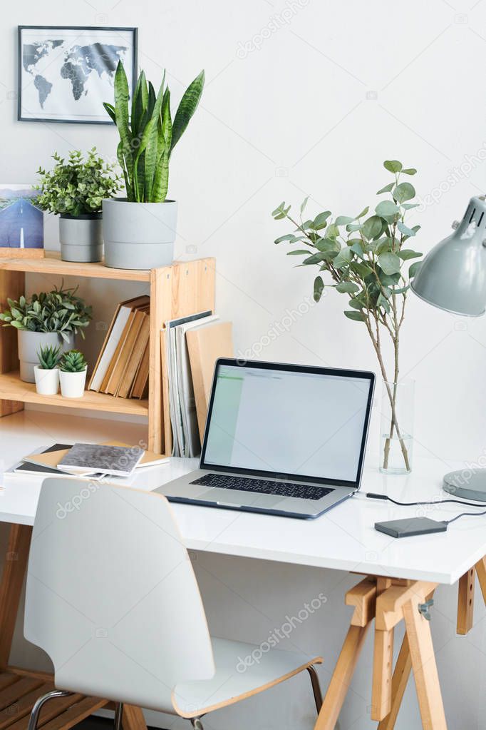 White chair by desk with lamp, laptop, books and copybooks and green plants in flowerpots on wooden shelves by wall forming workplace