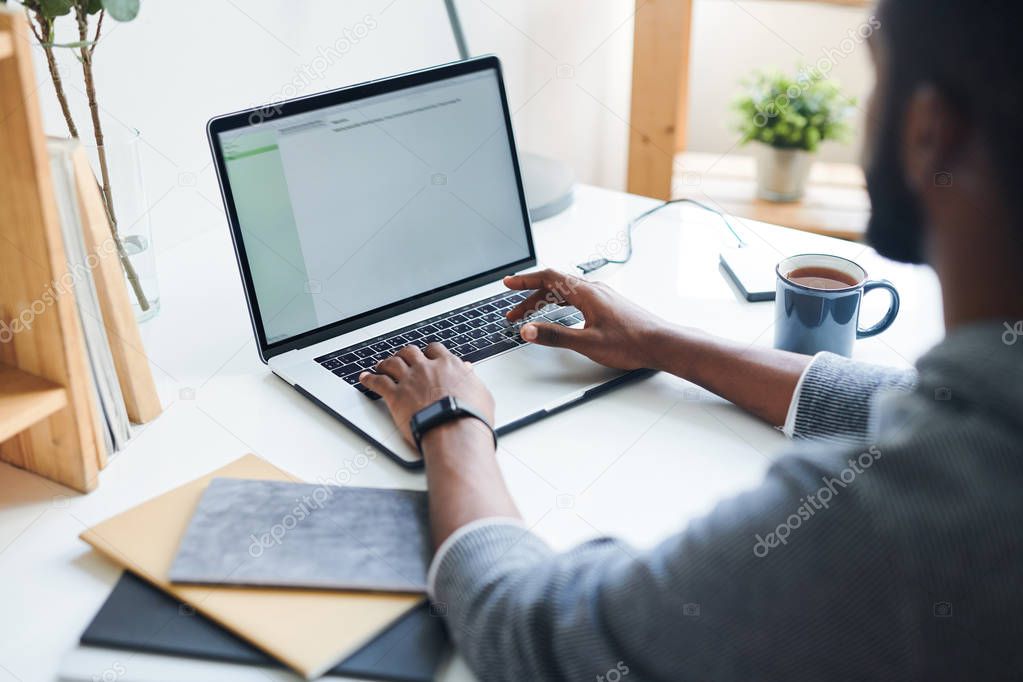 Multi-ethnic young employee or student typing in front of laptop while sitting by desk and preparing report for conference