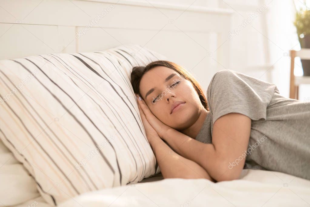 Pretty young brunette woman in grey t-shirt keeping her head on striped pillow while lying on bed and napping at home
