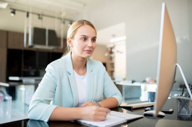 Genç zarif ofis çalışanı not alırken monitörün önünde otururken bilgisayar ekranına bakıyor