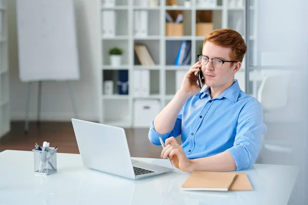 Young serious manager in blue shirt talking by smartphone while sitting by workplace in front of laptop