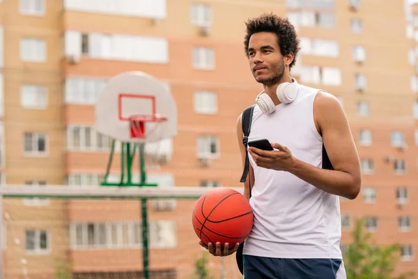 都市環境の遊び場で休憩時にボールとスマートフォンのテキストメッセージを持つ若い混合レースの選手 — ストック写真