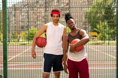 Kortu çevreleyen çitlerin yanında duran aktif giyimli iki genç profesyonel basketbolcudan oluşan küçük bir grup