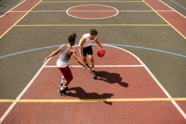 Genç basketbolcu oyun sırasında açık kort boyunca taşırken rakip topu savunmak için çalışıyor