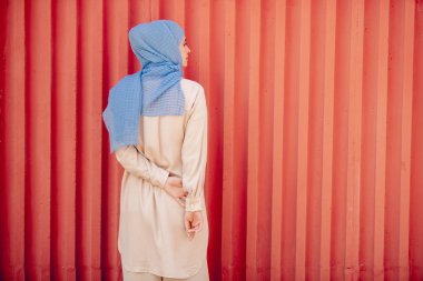 Mavi başörtüsü ve duvar başında duran açık bej casualwear genç Müslüman kadın geri görünümü