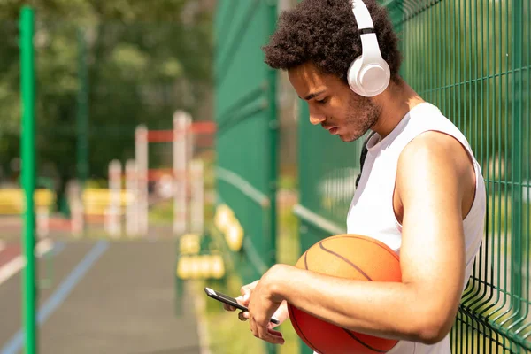 休憩時間にスマートフォンでスクロールしながらフェンスのそばに立つヘッドフォンの若い現代バスケットボール選手 — ストック写真