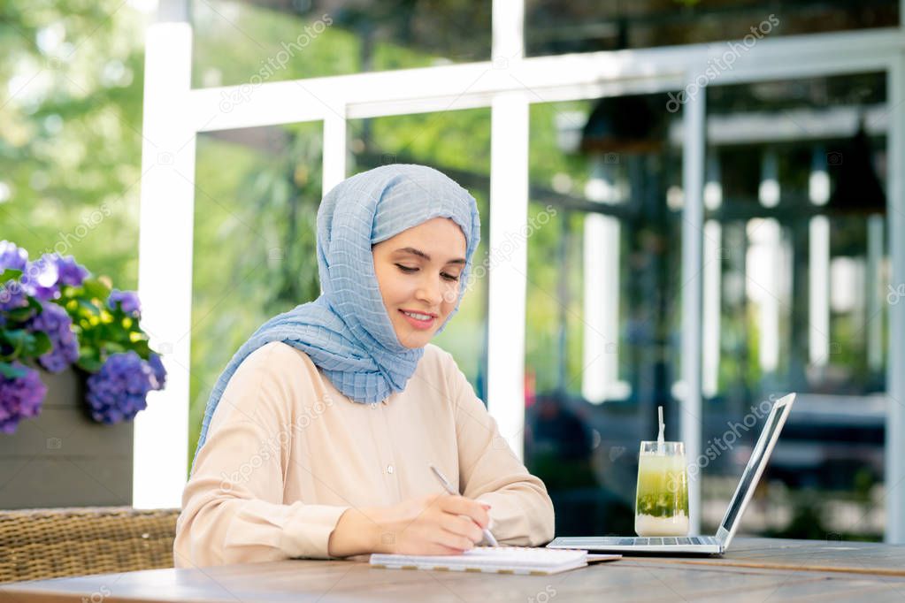 Young muslim student in hijab making working notes or doing home assignment outdoors