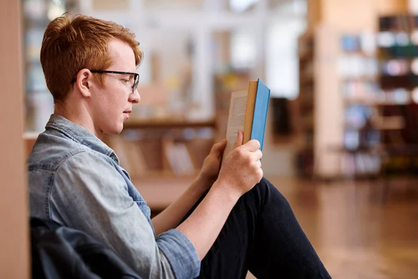 穿着休闲装的年轻读者坐在图书馆的书架旁 一边读有趣的书 — 图库照片