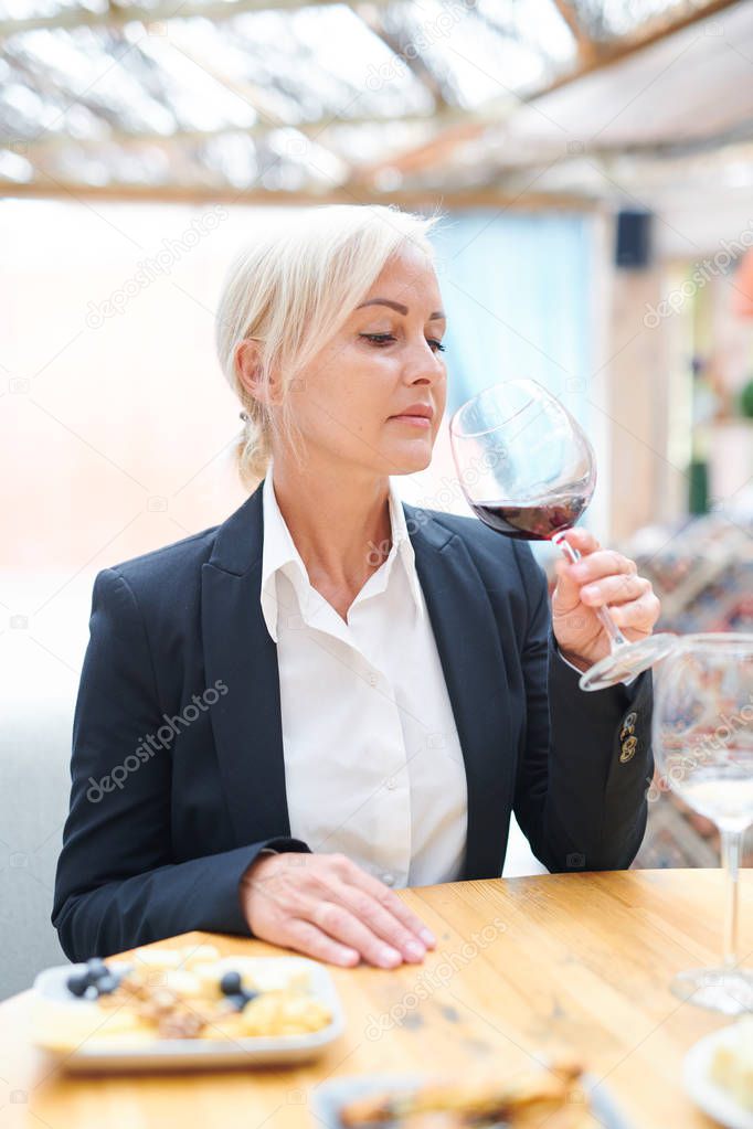 Professional female sommelier sitting by wooden table while evaluating smell and taste of red wine in restaurant