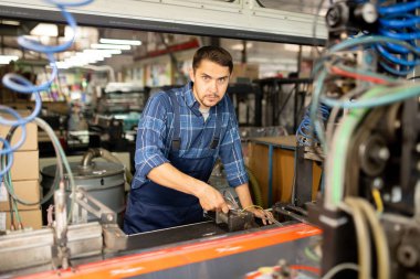 Endüstriyel makinelerden biriyle çalışan ve atölyede size bakan genç fabrika tamircisi veya teknisyeni