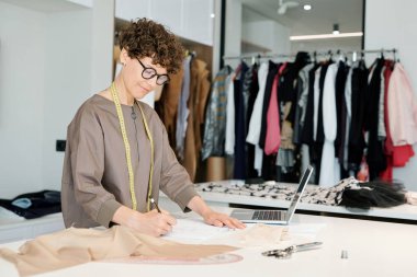 Pretty genç kadın terzi işyeri tarafından ayakta iken kağıt üzerinde yeni giysiler in kroki yapma