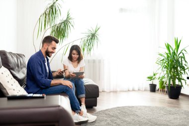 Wifi ile düz rahat kanepede oturan ve iletişim yerine cihazları kullanarak rahat kıyafetler içinde Pozitif genç çift