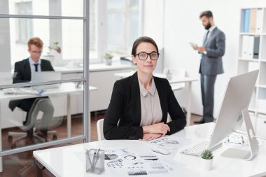 Ofiste, muhasebeci veya yönetici konseptinde masada otururken kameraya bakan gözlük takan genç iş kadınının portresi