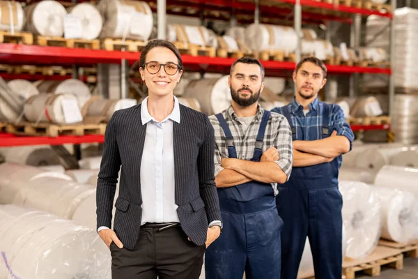 快乐年轻优雅的女领导工程队和两个穿着工作服的男子站在仓库内 — 图库照片