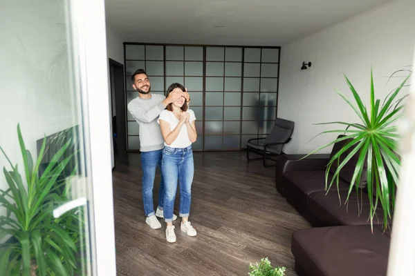 Rahat oturma odasında ayakta rahat kıyafetli gülümseyen yakışıklı genç sakallı adam ve onun için sürpriz hazırlarken eşine gözleri kaplayan