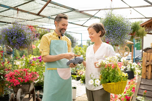 男性の庭師や販売員は バスケットでかわいい女性に鉢植えの花を販売し 非接触支払いアプリを使用してエプロンで — ストック写真