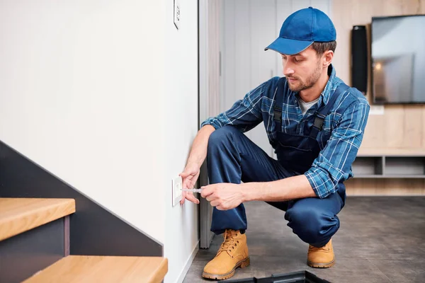 Young electrician from household maintenance service standing on squats by wall while checking voltage of socket