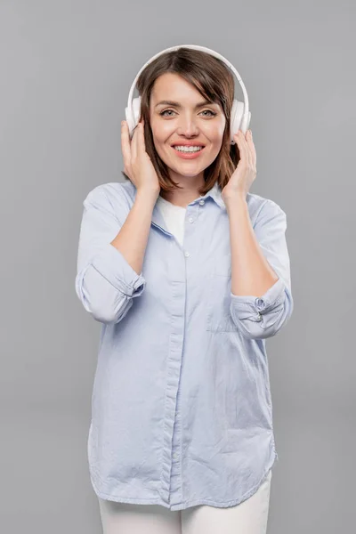 カジュアルな青いシャツと白いジーンズで笑顔の女の子は カメラの前でヘッドフォンで彼女のお気に入りの音楽を聴きます — ストック写真