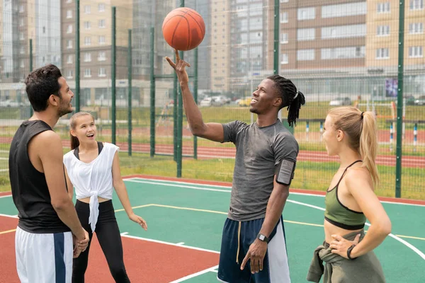 バスケットボールの遊び場で彼の驚くべき友人の間で人差し指にボールを保持する幸せな若いアフリカのスポーツマン — ストック写真
