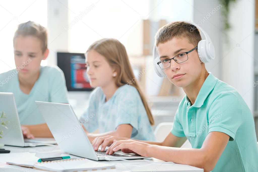Handsome schoolboy in headphones looking at you while typing on laptop keypad by desk with classmates on background