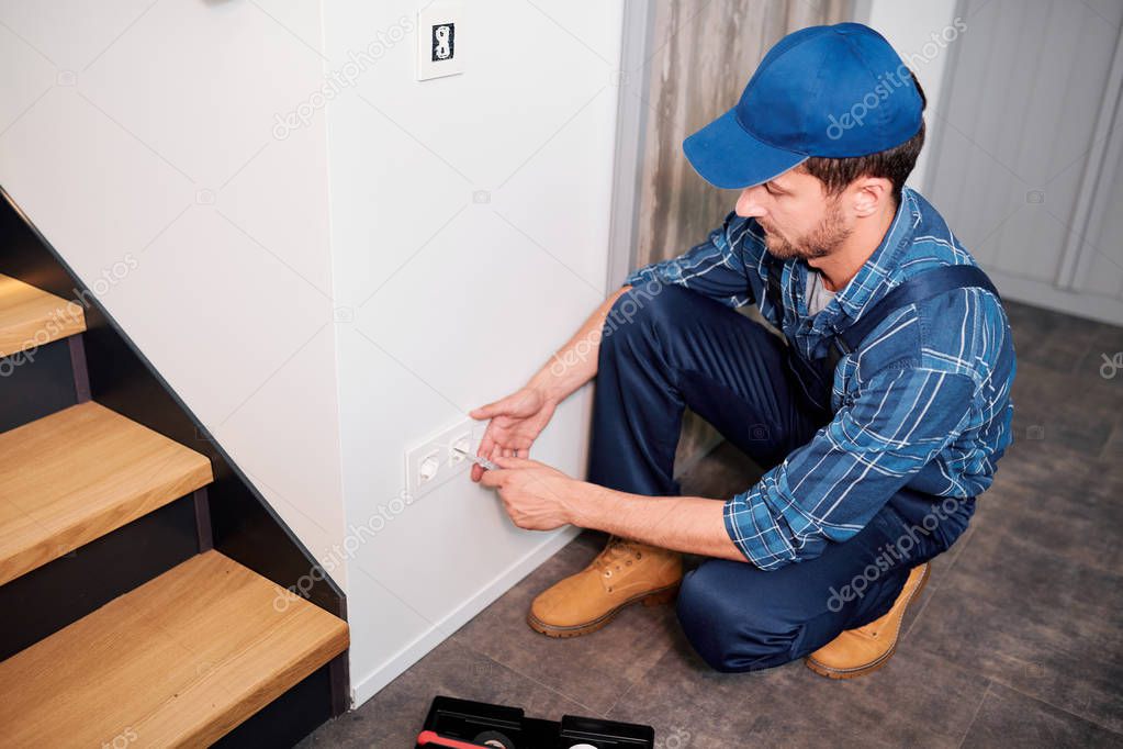 Professional electrician in workwear checking or measuring voltage of socket while sitting on squats by wall