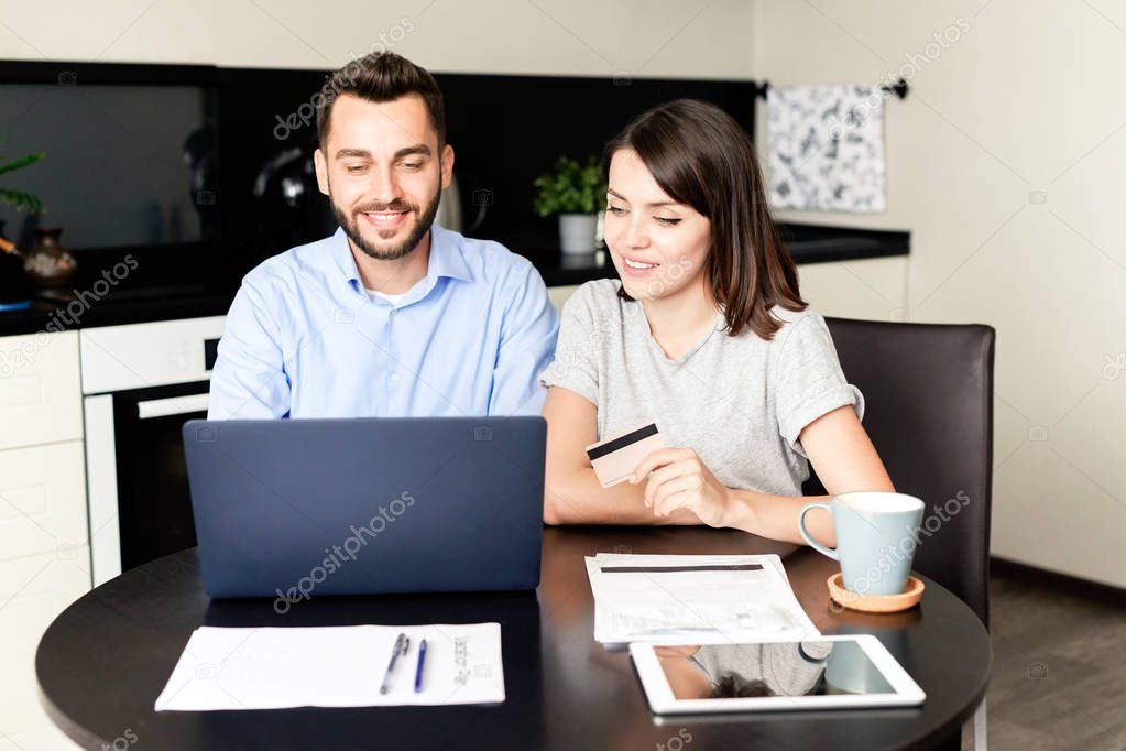Smiling young couple sitting at kitchen table and using laptop and credit card while paying bills online