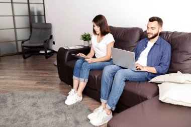 Genç çift evde cihazlar ile meşgul: erkek ve kadın deri kanepede oturan ve cihazlarda net sörf