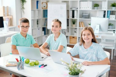 Sınıfta dizüstü bilgisayarların önünde masa başında otururken size bakan üç çalışkan öğrenci