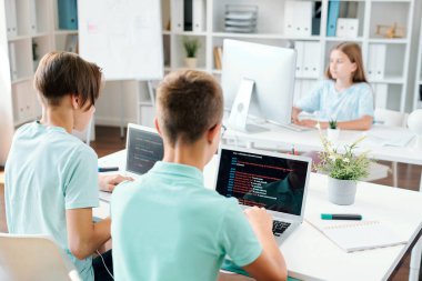 Dizüstü bilgisayarlarla oturan, ekranlarla ilgili verilere bakan ve okul projesi üzerinde tek tek çalışan iki genç çocuk