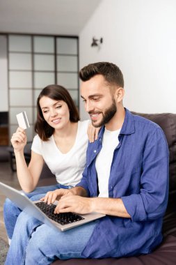 Neşeli genç çift oturma odasında kanepede oturan ve sipariş için online ödeme yaparken kredi kartı ve dizüstü bilgisayar kullanarak