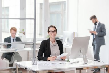 Ofiste masada otururken kameraya bakan gözlük takan genç iş kadınının portresi, stajyer veya yönetici kavramı