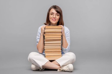 Genç zeki öğrenci önünde yüksek kitap yığını ile katta oturan ve size bakıyor