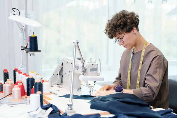 漂亮的年轻裁缝坐在缝纫机和加工蓝色纺织品 而工作在一件衣服 — 图库照片