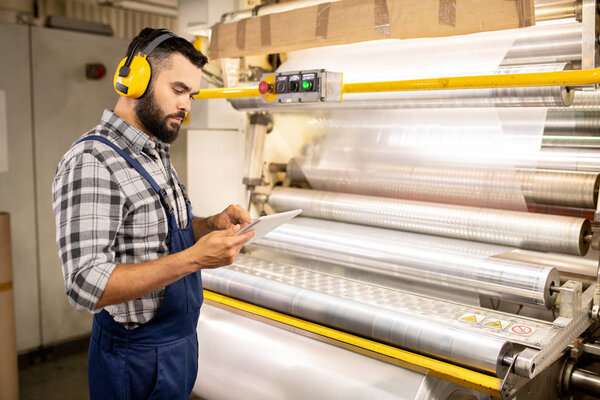 Молодой серьезный инженер по контролю качества с помощью планшета для проверки параметров обрабатываемого материала при нахождении на производственной машине
