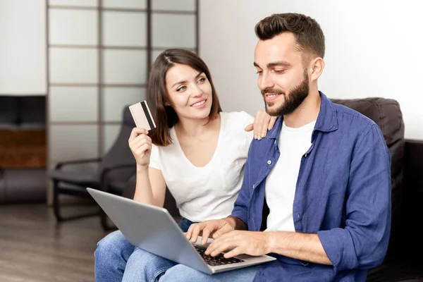 Positive beautiful couple in casual clothing sitting on sofa at home and using laptop while buying furniture on internet