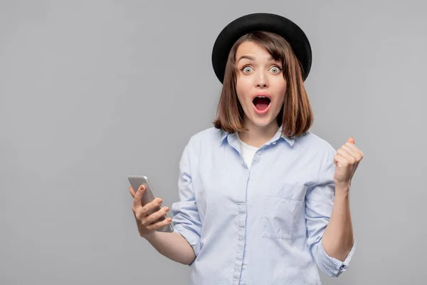 相当惊讶的女孩在帽子和衬衫看着你 而拿着智能手机与促销销售 — 图库照片